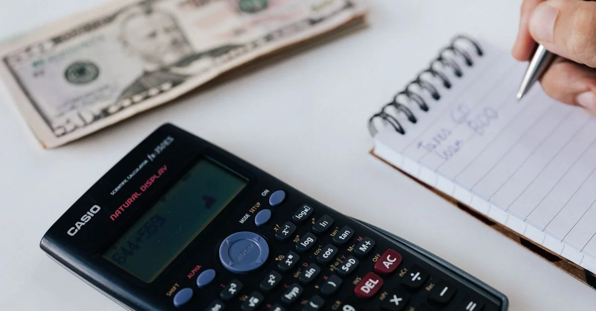 Gros plan sur une calculatrice, un billet en dollars américains et une main écrivant dans un carnet, symbolisant la planification financière.